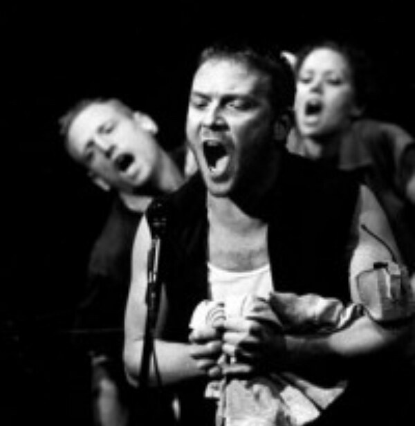 Ari Fliakos performing onstage with two other performers, mid-shout at a microphone